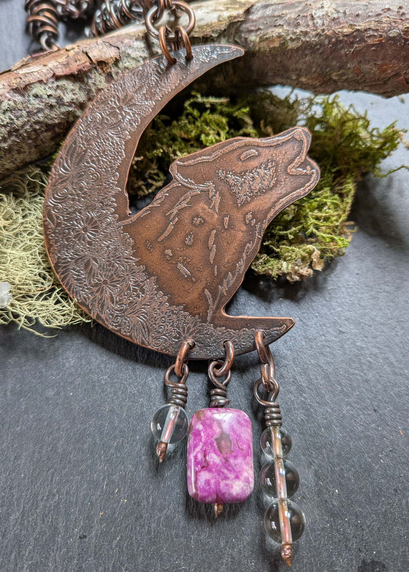 A handcrafted copper wolf and moon pendant resting on warm cream linen and weathered oak with moss and heather